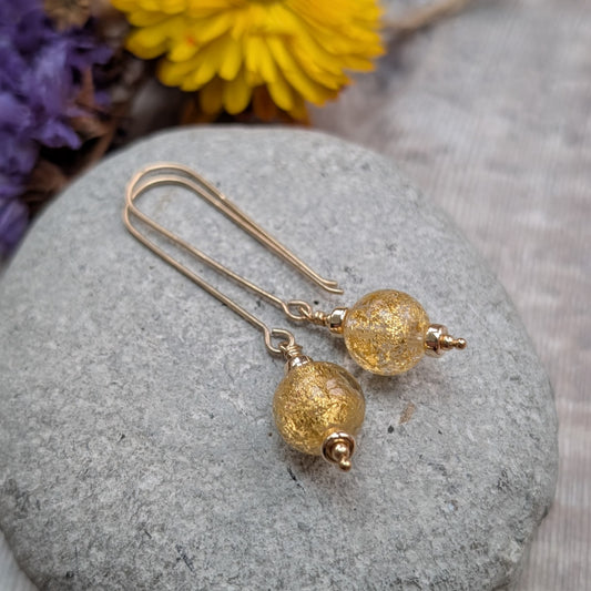 Pair of long gold filled drop earrings with round gold fleck Venetian glass beads, resting on a smooth grey stone with dried flowers in the background.