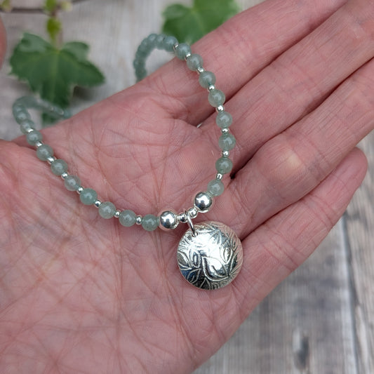 Green Aventurine and Silver Leaf Disc Necklace held in the palm of a hand, showing the size of the textured silver disc charm and the pale green gemstone beads.