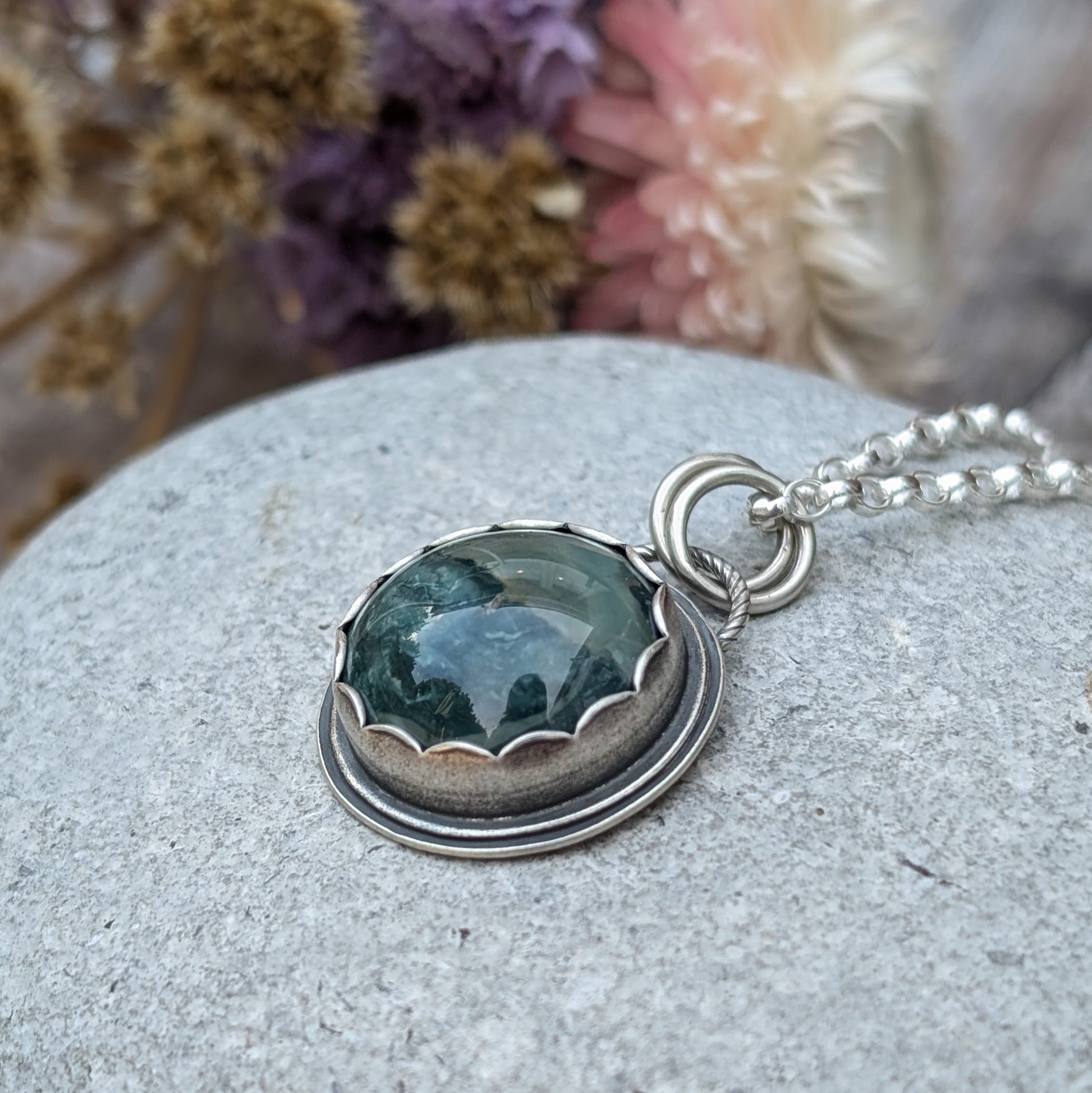 A close-up image of a handcrafted necklace featuring a Green Hava Ridge Jasper stone set in a recycled silver bezel, laid on a smooth grey stone surface.
