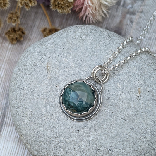 A close-up image of a handcrafted necklace featuring a Green Hava Ridge Jasper stone set in a recycled silver bezel, laid on a smooth grey stone surface.