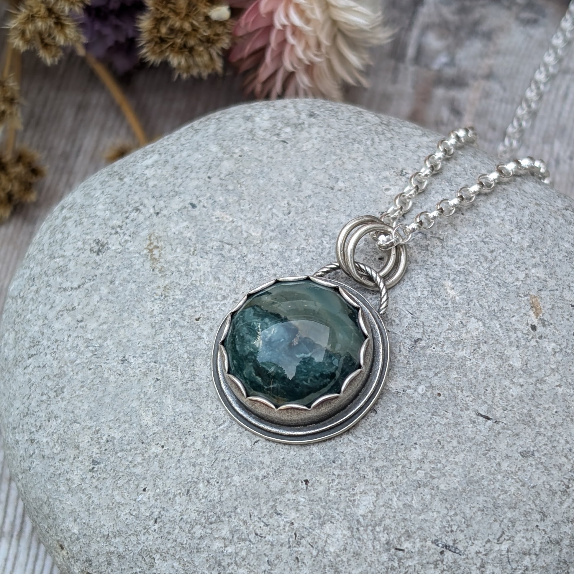 A close-up image of a handcrafted necklace featuring a Green Hava Ridge Jasper stone set in a recycled silver bezel, laid on a smooth grey stone surface.
