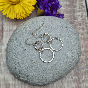 Sterling Silver two circle drop earrings. Each earring has one smaller silver open circle attached to silver earring wire. Dropping from the first open circle via two small silver hoops is a larger silver open circle. Each circle has slightly hammered texture, the largest circle measuring approximately 15mm diameter. Total drop from earlobe approximately 35mm providing lots of movement.