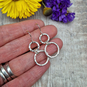 Sterling Silver two circle drop earrings. Each earring has one smaller silver open circle attached to silver earring wire. Dropping from the first open circle via two small silver hoops is a larger silver open circle. Each circle has slightly hammered texture, the largest circle measuring approximately 15mm diameter. Total drop from earlobe approximately 35mm providing lots of movement.