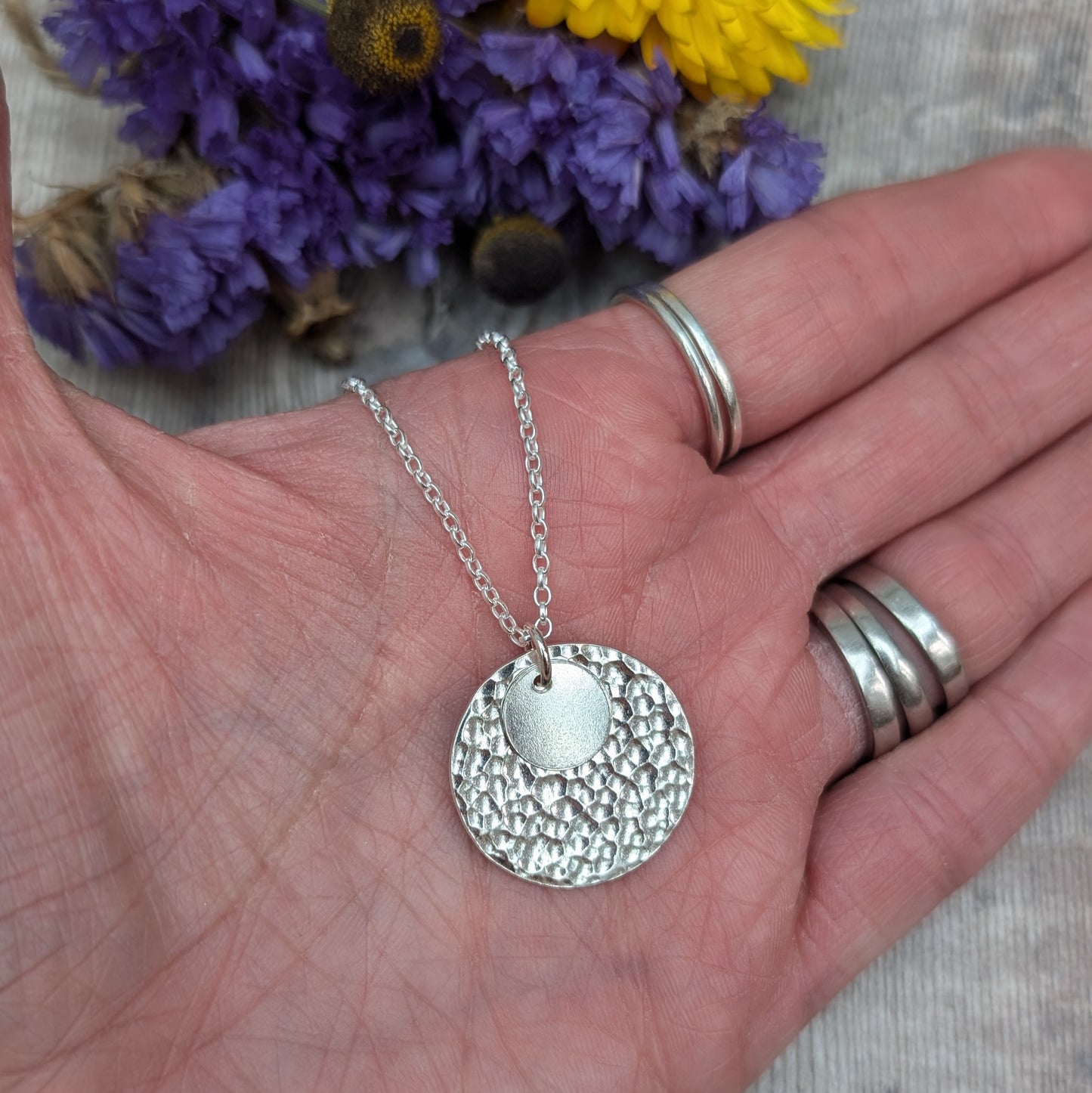 Close-up of a hand holding a handmade silver necklace with two hammered discs, one small and one large, with blurred purple and yellow flowers in the background.