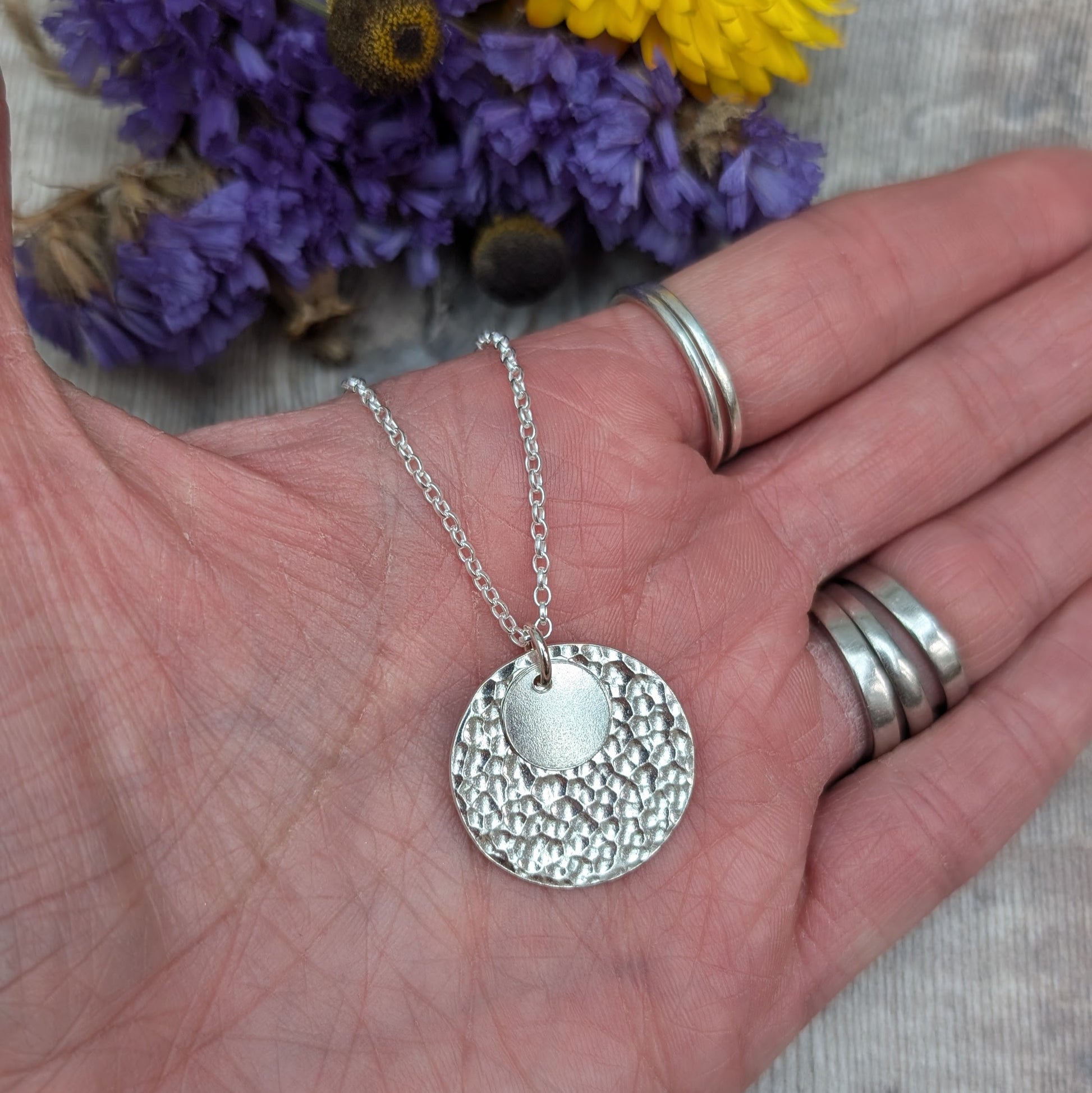 Close-up of a hand holding a handmade silver necklace with two hammered discs, one small and one large, with blurred purple and yellow flowers in the background.