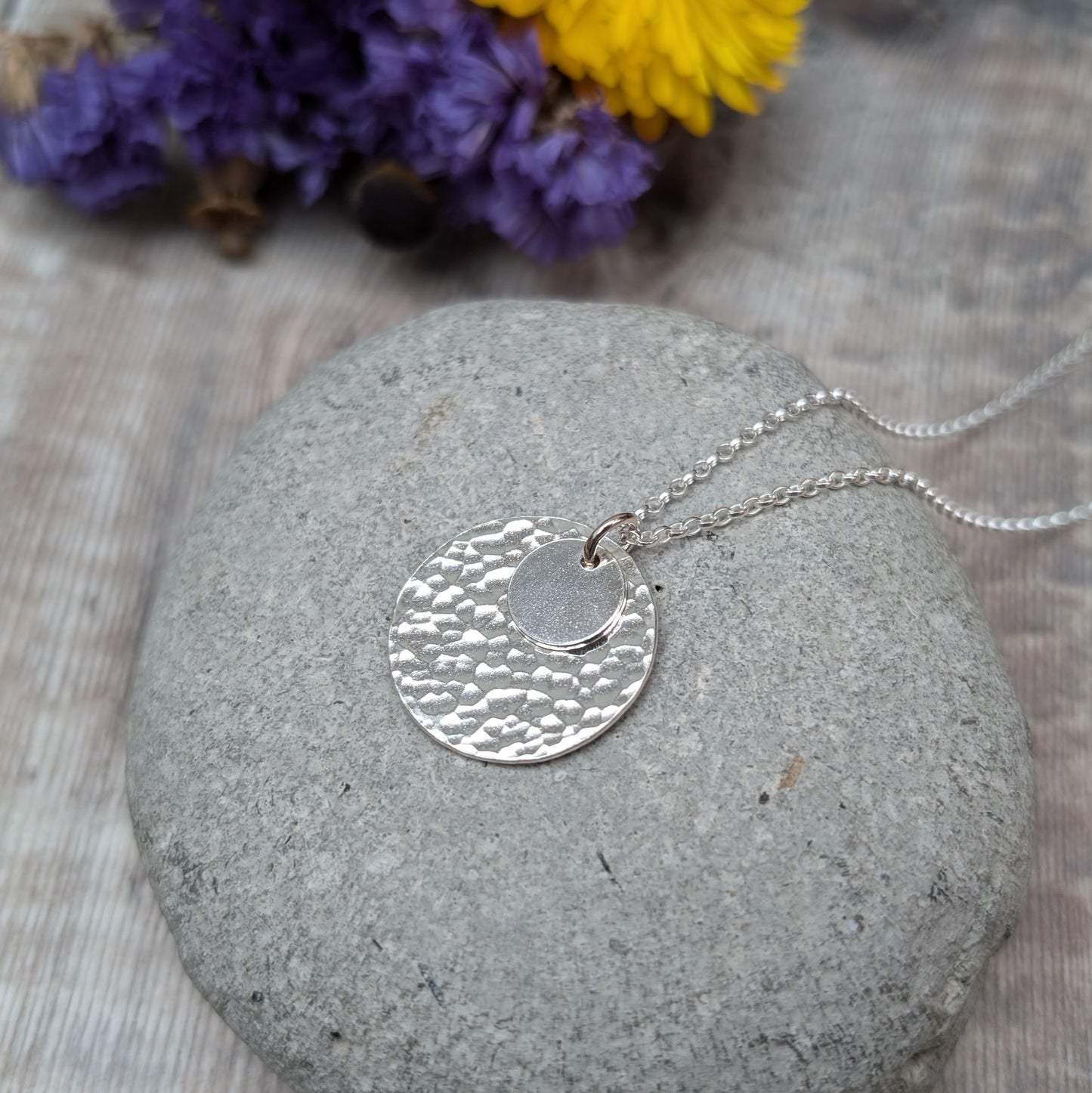 Handmade silver necklace featuring two hammered discs, one small and one large, displayed on a smooth grey stone with blurred purple and yellow flowers in the background.