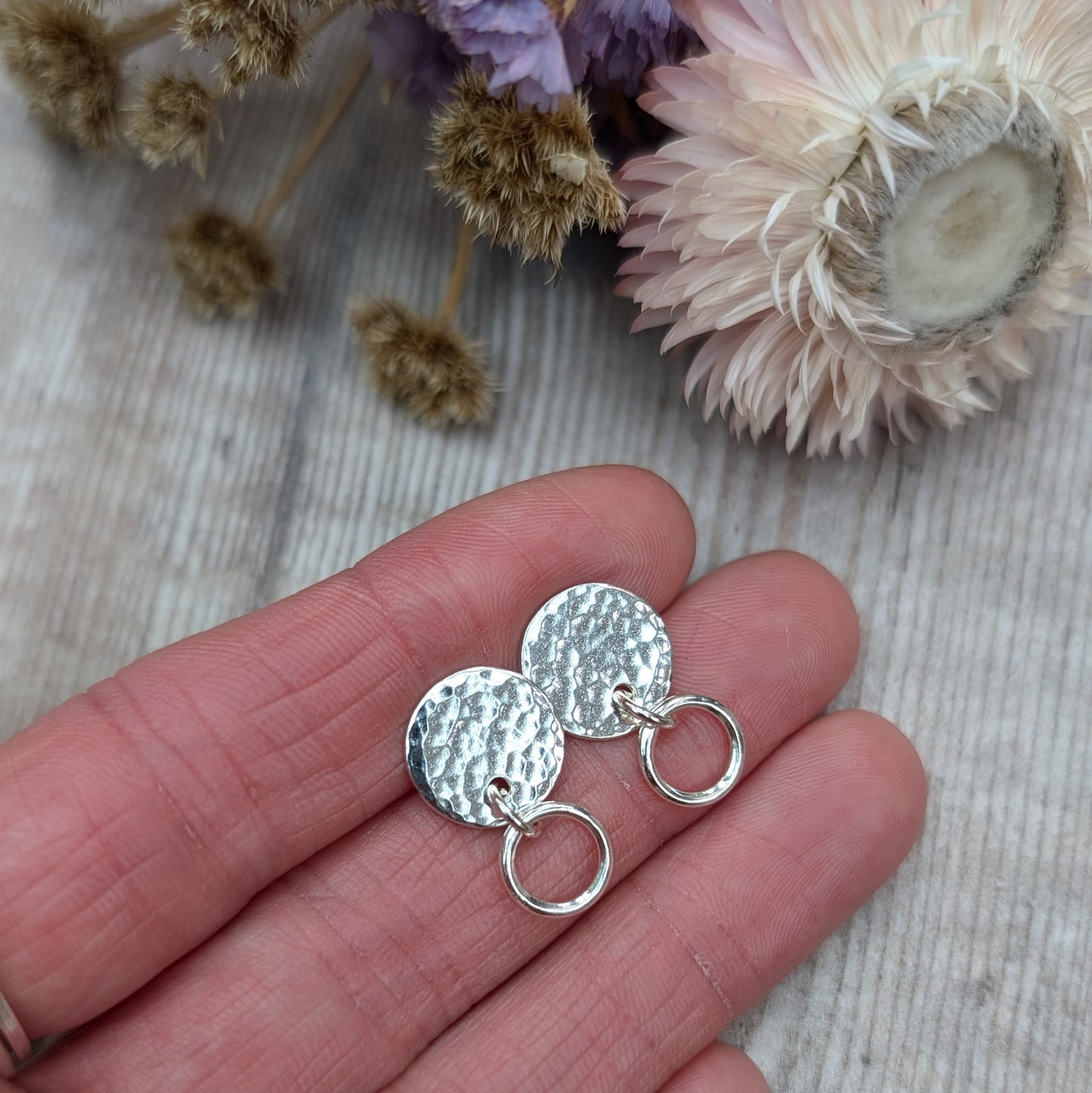 Close-up of a hand holding a pair of handmade stud earrings with a hammered silver disc and a small hoop, placed against a light wooden background with soft-focus dried flowers in the background.
