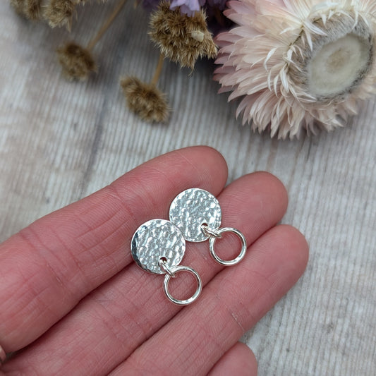 Close-up of a hand holding a pair of handmade stud earrings with a hammered silver disc and a small hoop, placed against a light wooden background with soft-focus dried flowers in the background.
