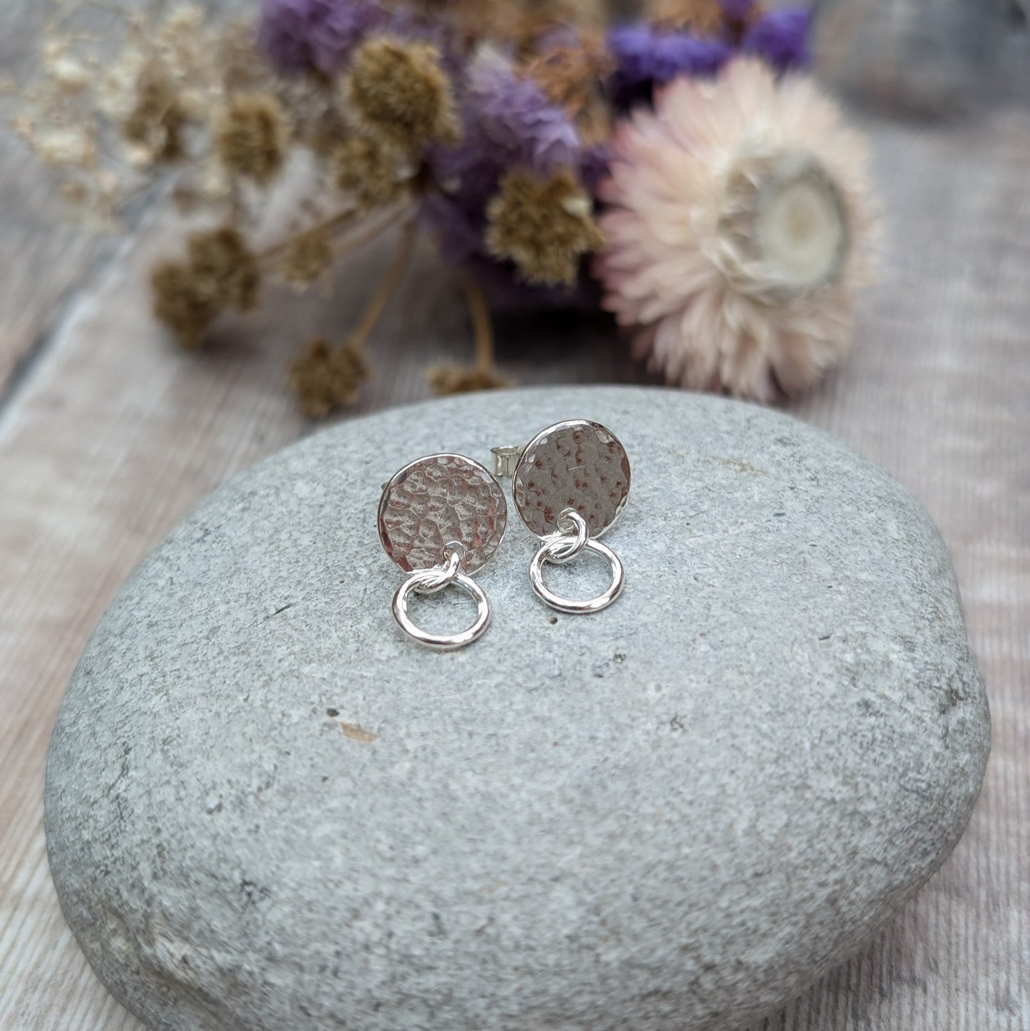 A pair of handmade stud earrings featuring a hammered silver disc with a small hoop, displayed on a smooth grey stone with purple and beige dried flowers softly blurred in the background
