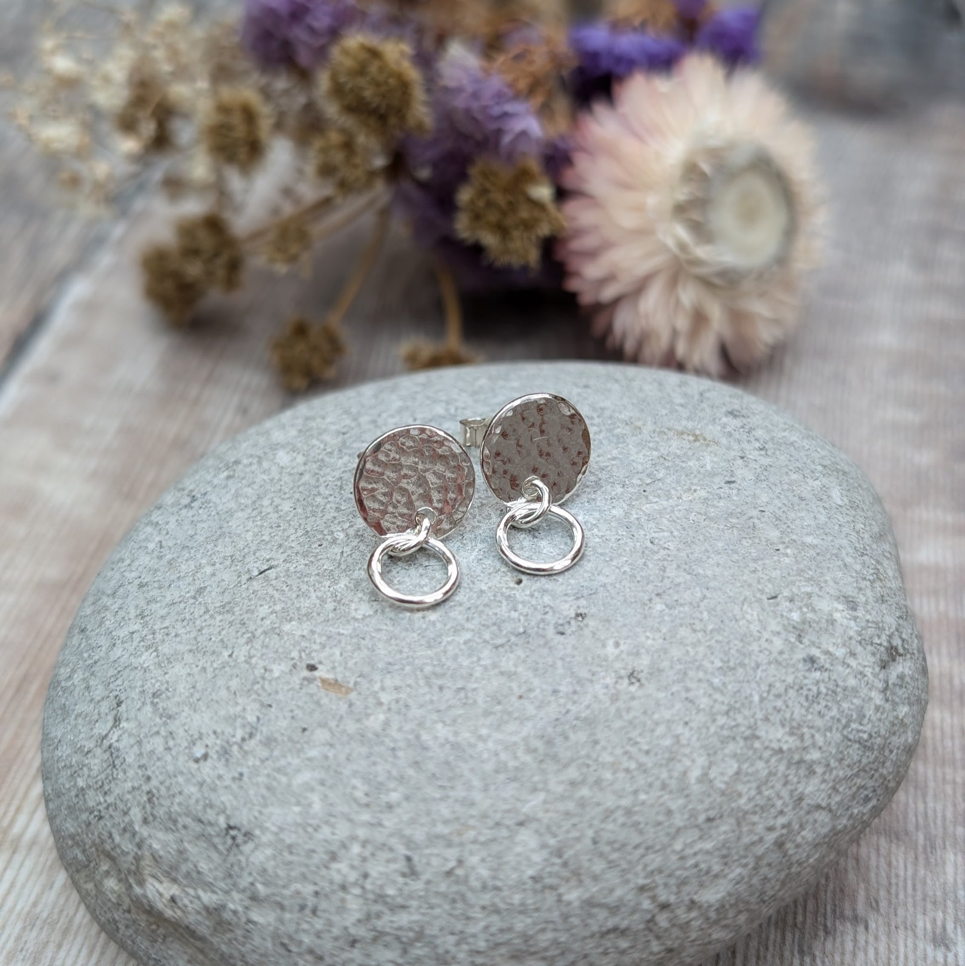 A pair of handmade stud earrings featuring a hammered silver disc with a small hoop, displayed on a smooth grey stone with purple and beige dried flowers softly blurred in the background