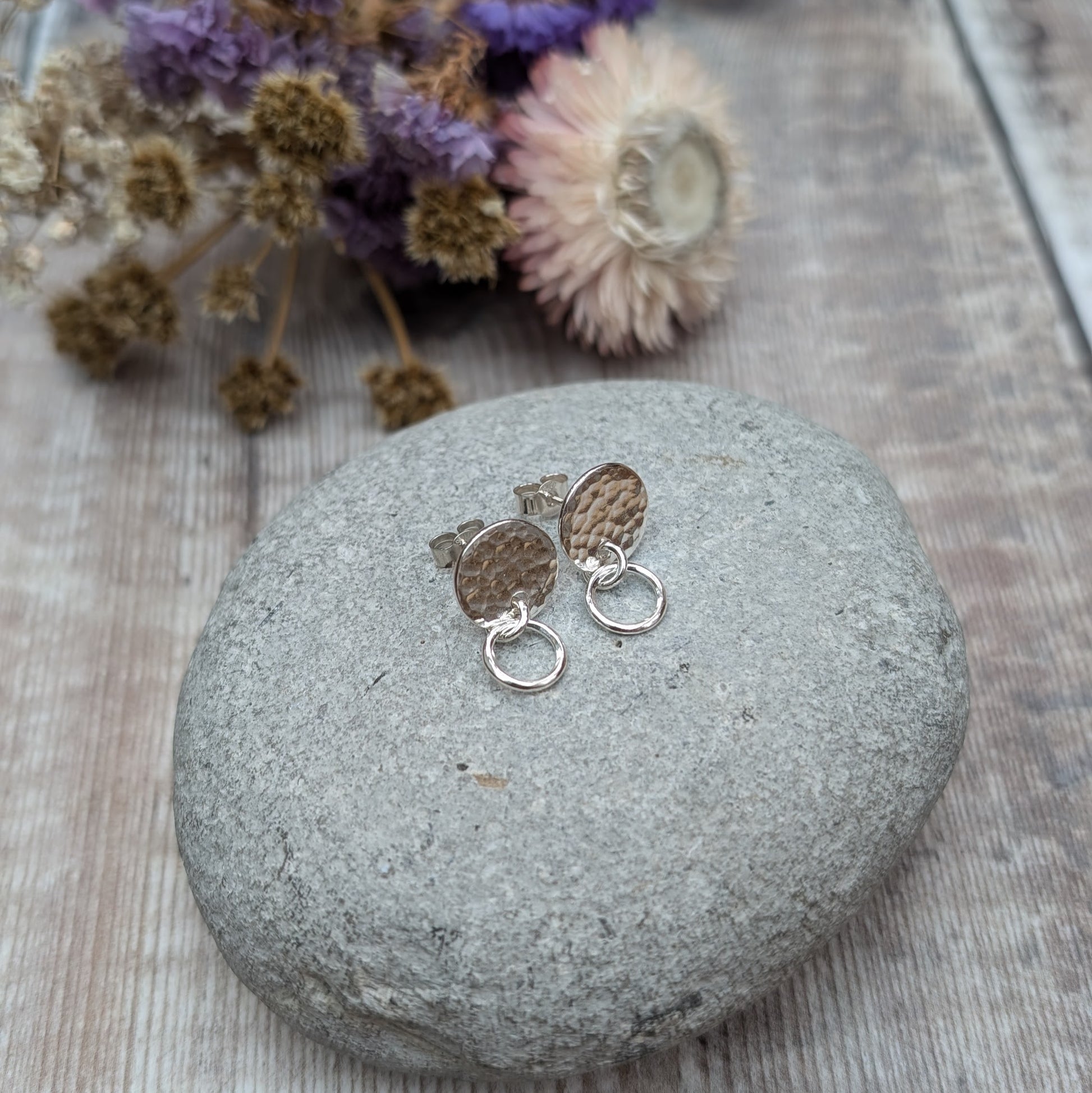A pair of handmade stud earrings featuring a hammered silver disc with a small hoop, displayed on a smooth grey stone with purple and beige dried flowers softly blurred in the background