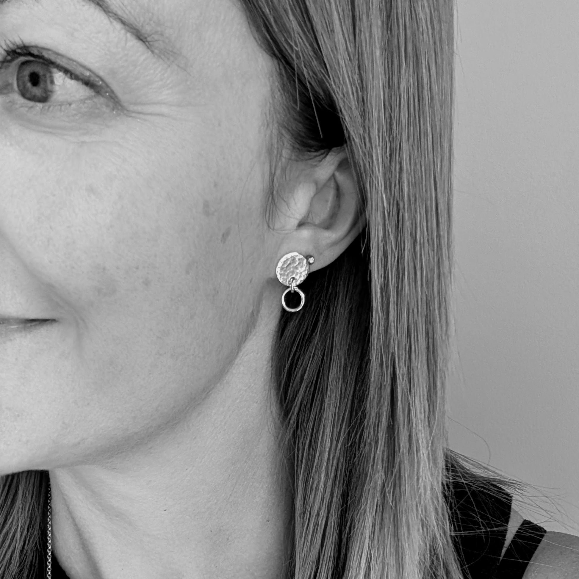 Black and white image showing a woman wearing a handmade stud earring with a hammered silver disc and a small hoop, highlighting the earring on the left ear with a neutral background.