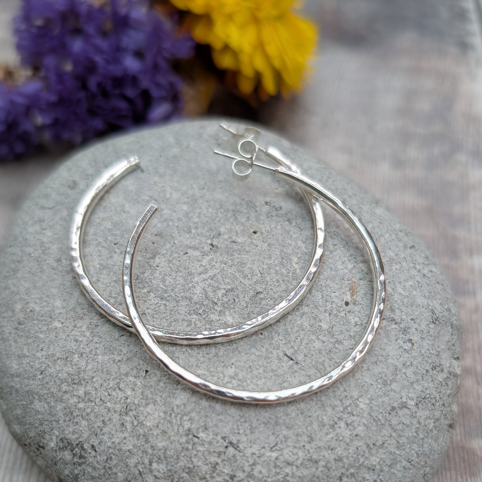 Hammered sterling silver hoop earrings arranged on a smooth grey stone, with purple and yellow flowers in the background.