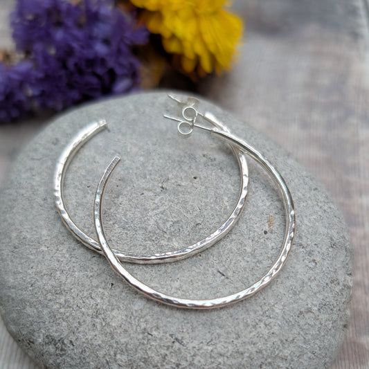 Hammered sterling silver hoop earrings arranged on a smooth grey stone, with purple and yellow flowers in the background.