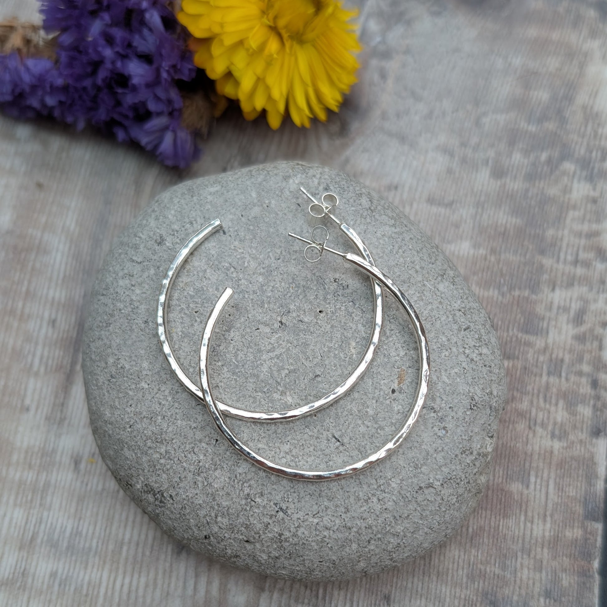 Hammered sterling silver hoop earrings arranged on a smooth grey stone, with purple and yellow flowers in the background.