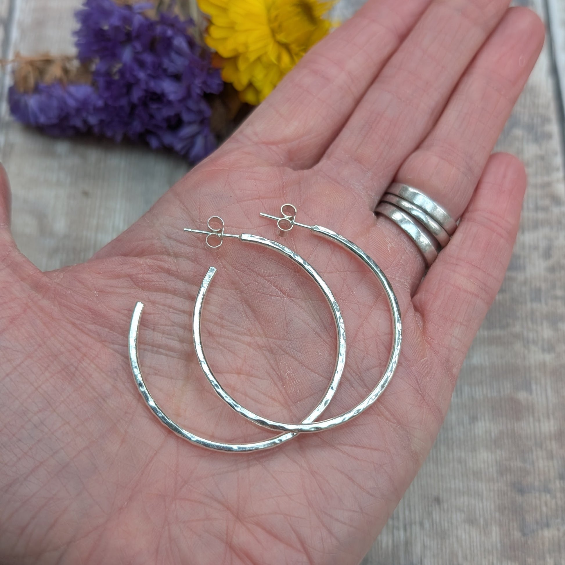 A pair of large 40mm hammered silver hoop earrings displayed on a hand for scale, with a blurred background of yellow and purple flowers.