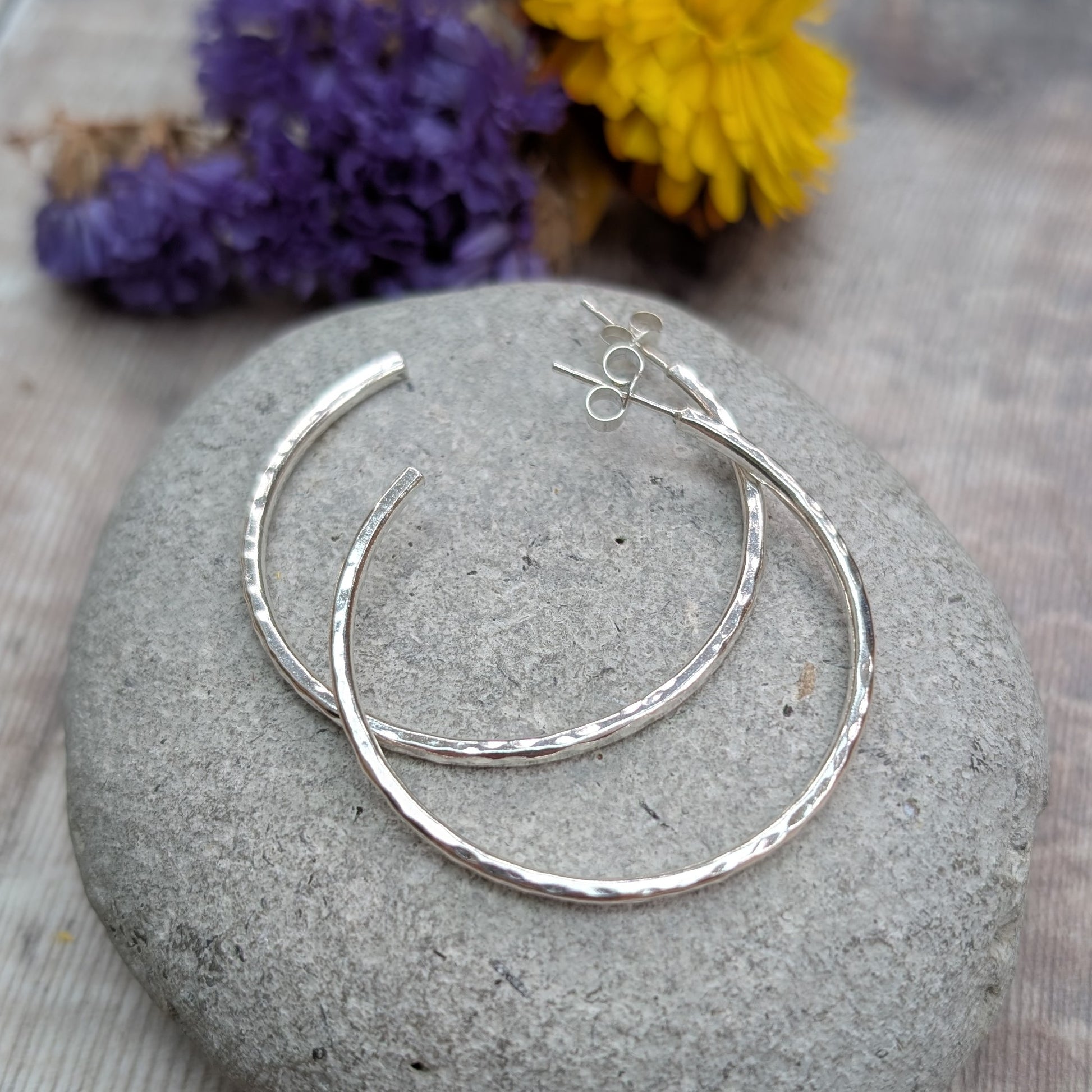 Hammered sterling silver hoop earrings arranged on a smooth grey stone, with purple and yellow flowers in the background.