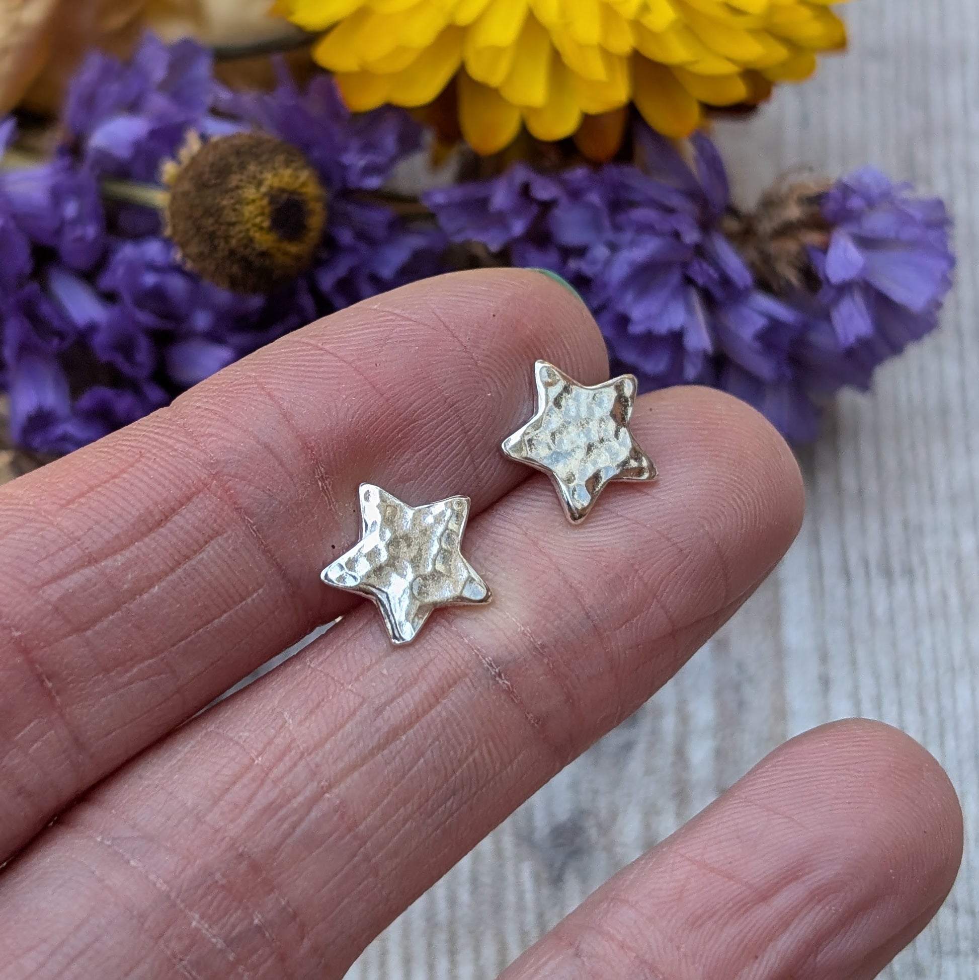 Close-up of a pair of handmade sterling silver star-shaped stud earrings with a hammered texture, displayed on the fingertips of a hand, with purple and yellow flowers in the background.