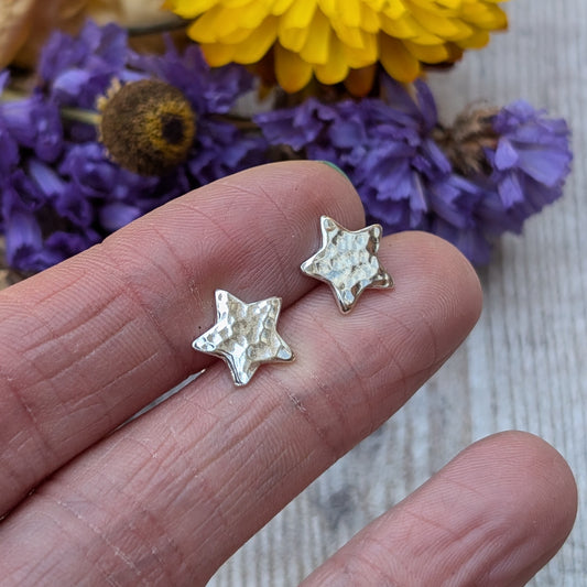 Close-up of a pair of handmade sterling silver star-shaped stud earrings with a hammered texture, displayed on the fingertips of a hand, with purple and yellow flowers in the background.