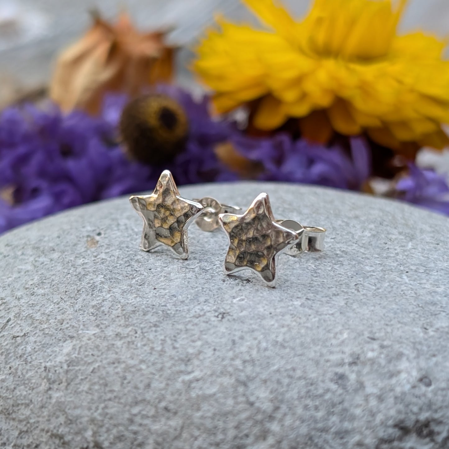 Close-up of a pair of handmade sterling silver star-shaped stud earrings with a hammered texture, resting on a smooth grey stone with vibrant yellow and purple flowers in the background.