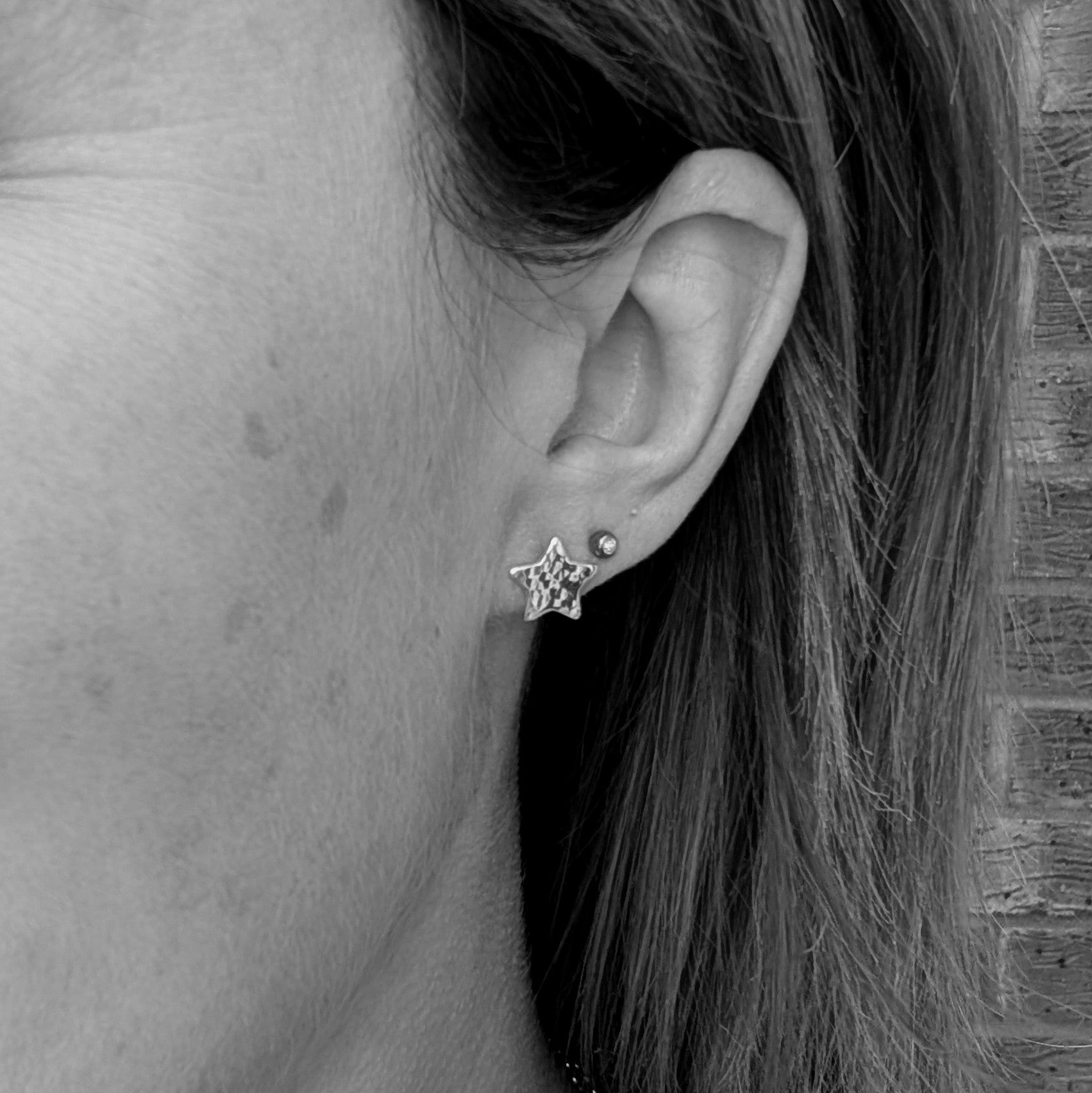 Black and white close-up of a person wearing a handmade sterling silver star-shaped stud earring with a hammered finish, alongside a smaller second stud in the ear.