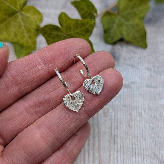 Close up of silver heart drop hoop earrings handmade from recycled Sterling Silver held in the hand showing the floral textured heart charms.