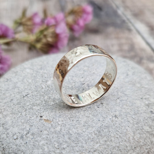 Handmade hammered ring with a polished finish, resting on a smooth stone. The ring has a subtle hidden message engraved on the inside. Background includes soft purple flowers."