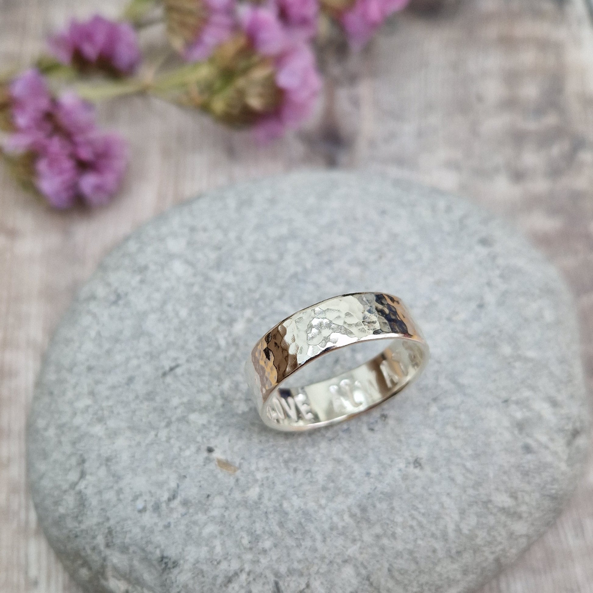 Handmade hammered ring with a polished finish, resting on a smooth stone. The ring has a subtle hidden message engraved on the inside. Background includes soft purple flowers."