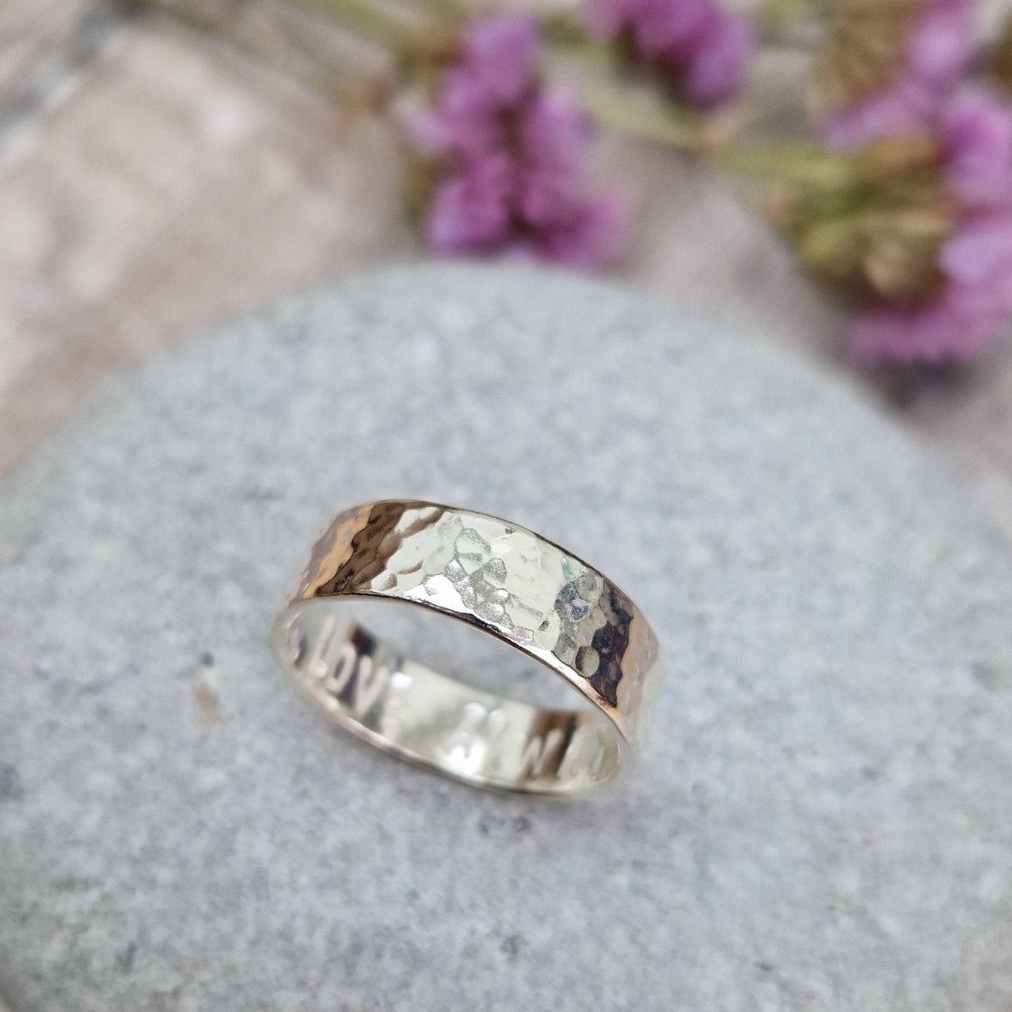 Handmade hammered ring with a polished finish, resting on a smooth stone. The ring has a subtle hidden message engraved on the inside. Background includes soft purple flowers."