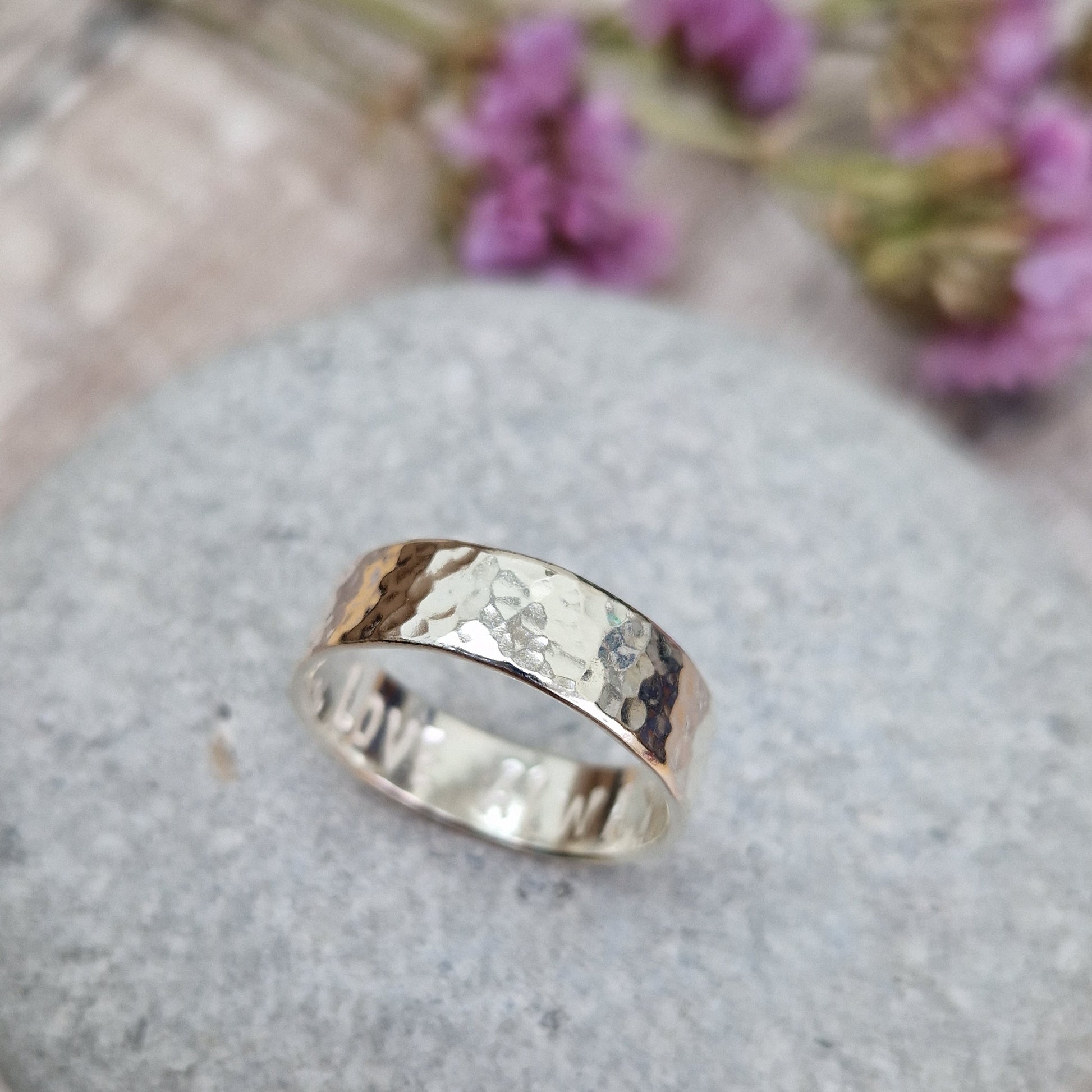 Handmade hammered ring with a polished finish, resting on a smooth stone. The ring has a subtle hidden message engraved on the inside. Background includes soft purple flowers."