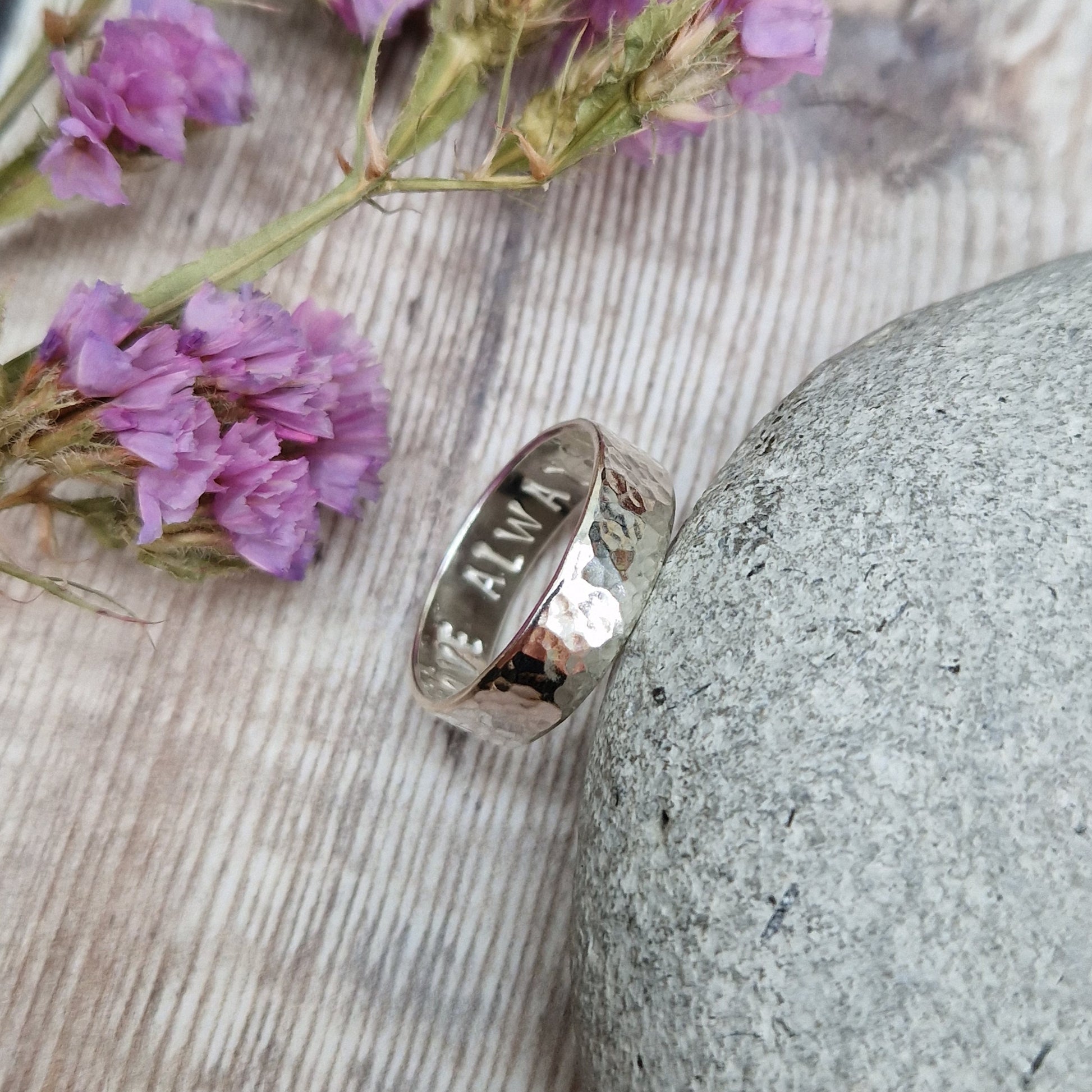 Close-up of a handmade hammered ring leaning against a stone. The polished surface reveals an engraved message on the inside. The background features rustic wood and purple flowers