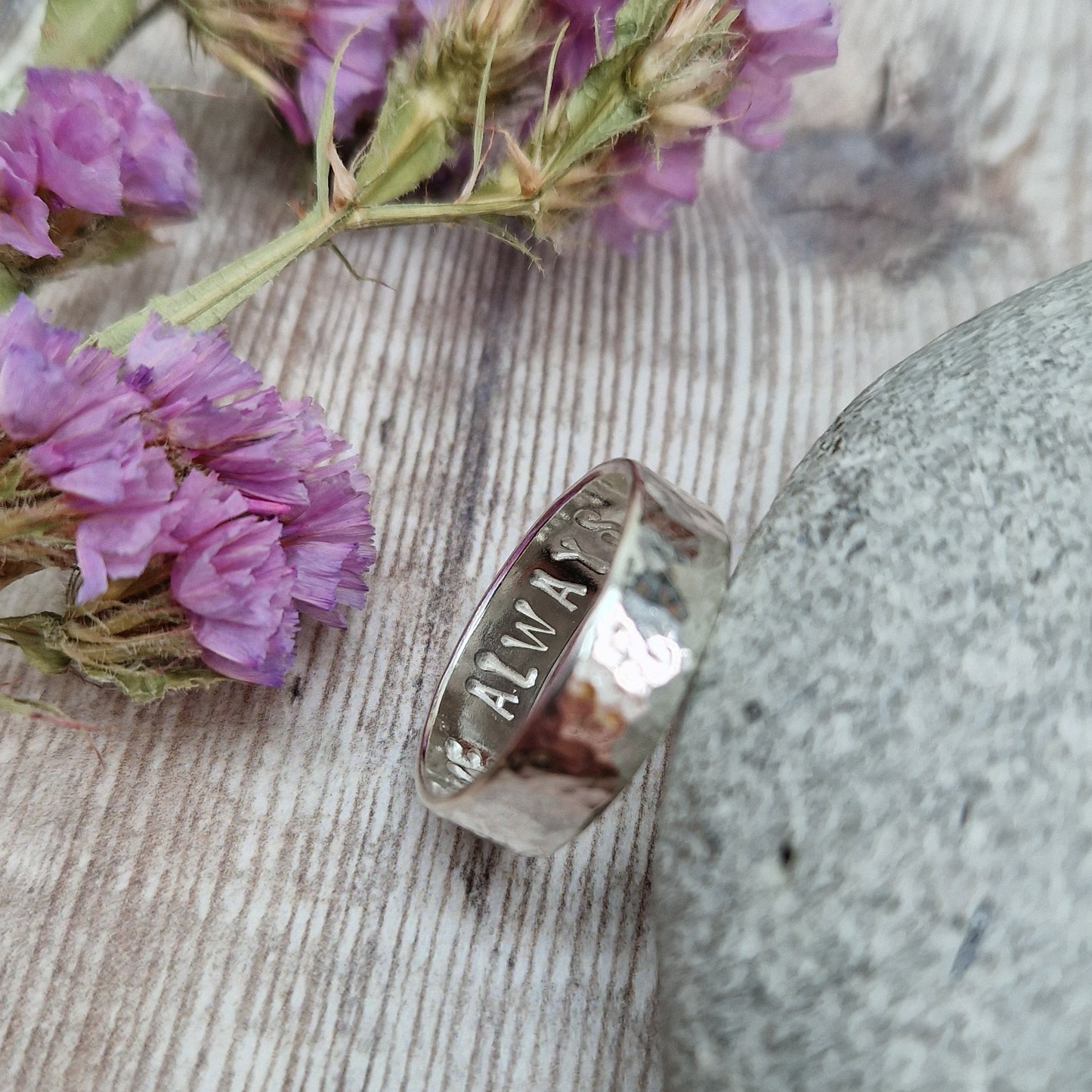 Close-up of a handmade hammered ring leaning against a stone. The polished surface reveals an engraved message on the inside. The background features rustic wood and purple flowers