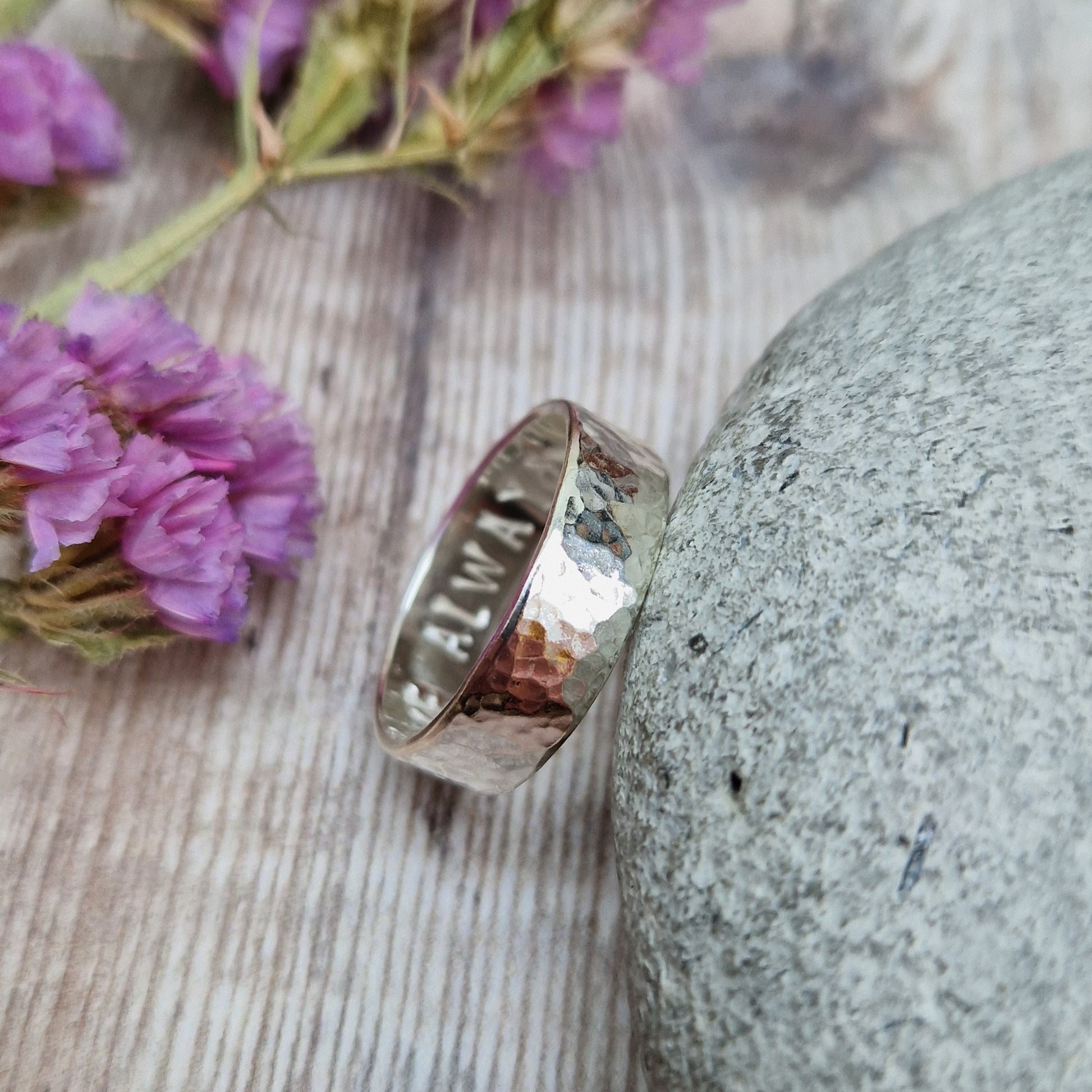 Close-up of a handmade hammered ring leaning against a stone. The polished surface reveals an engraved message on the inside. The background features rustic wood and purple flowers
