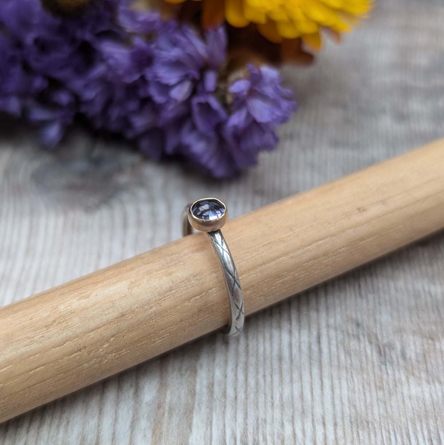 Silver ring with an iolite stone in a gold bezel, displayed on a wooden dowel. The band features an etched pattern, and the background includes purple flowers, adding a natural touch to the setting.