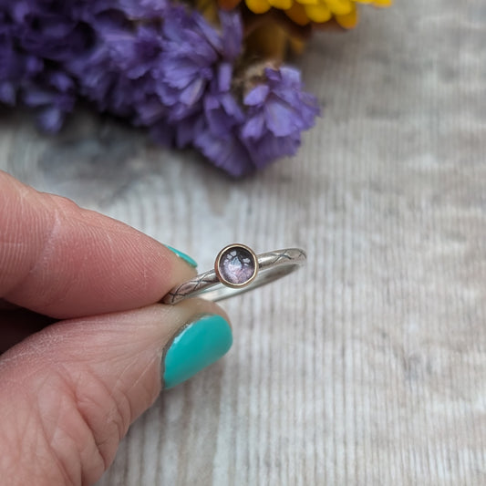 Hand holding a silver ring with a delicate iolite stone set in a gold bezel. The band has a subtle etched pattern, and the background features purple flowers for a touch of color.