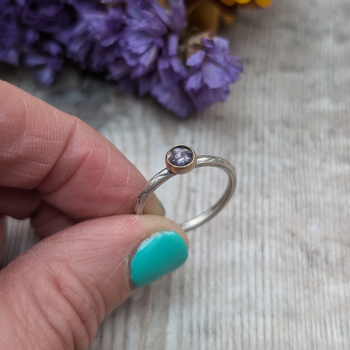 Hand holding a silver ring with a delicate iolite stone set in a gold bezel. The band has a subtle etched pattern, and the background features purple flowers for a touch of color.