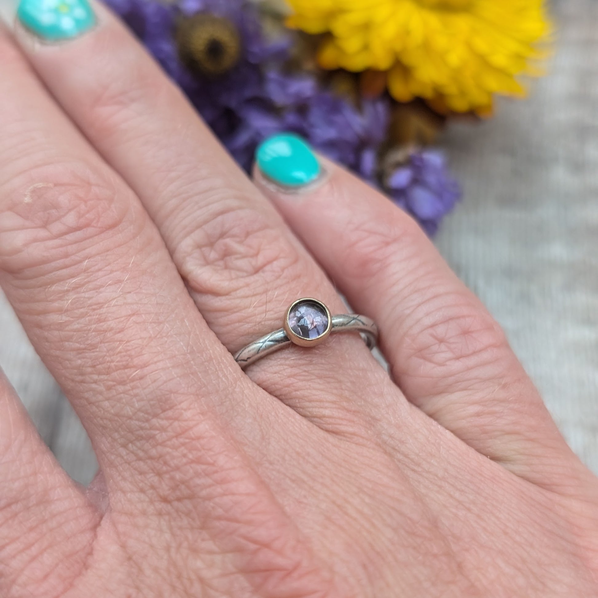 Silver ring with an iolite gemstone set in a gold bezel, worn on a finger. The etched pattern on the band and the deep purple hue of the stone create an elegant and unique look. Background shows blurred flowers in purple and yellow.