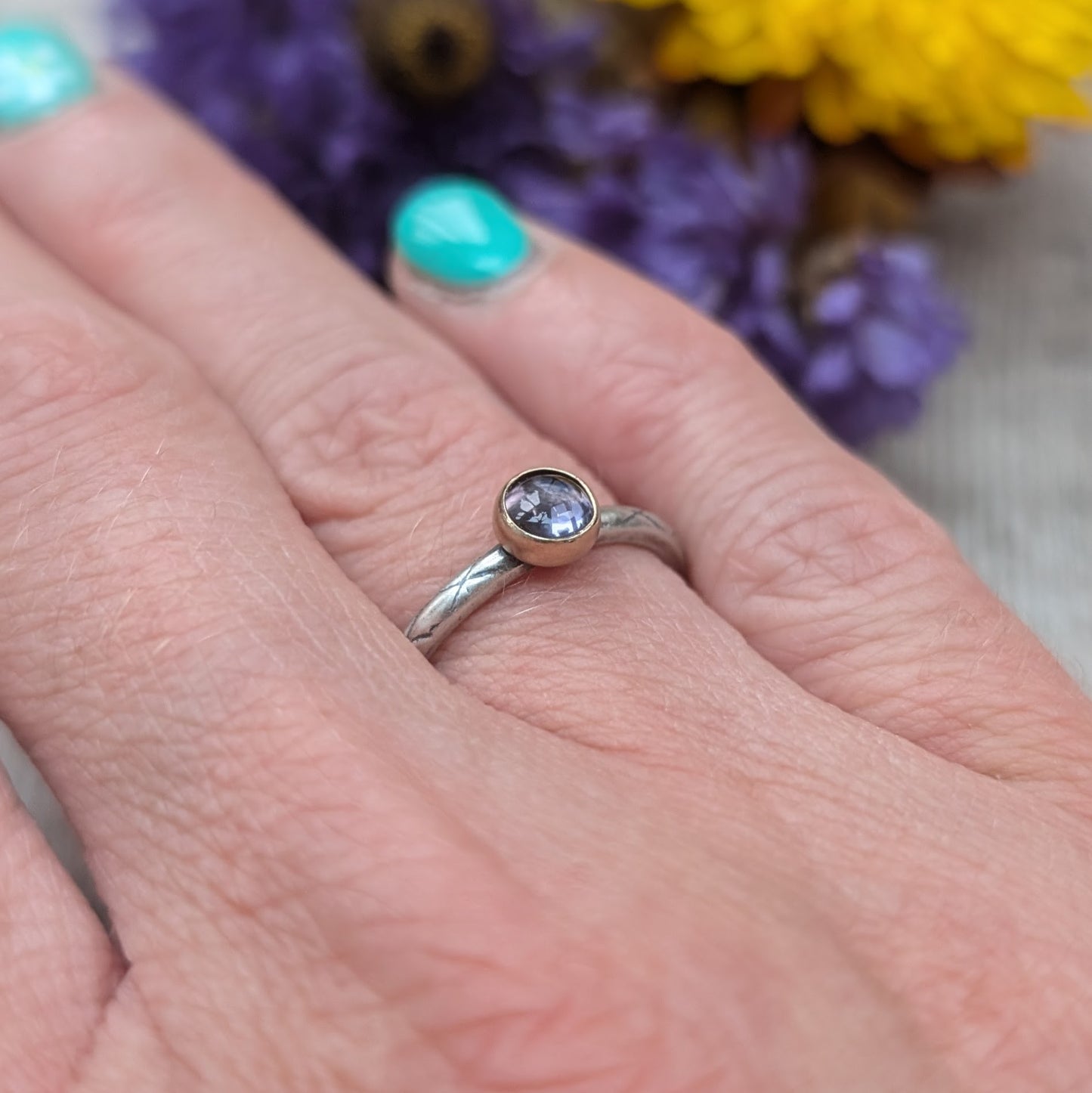 Silver ring with an iolite gemstone set in a gold bezel, worn on a finger. The etched pattern on the band and the deep purple hue of the stone create an elegant and unique look. Background shows blurred flowers in purple and yellow.