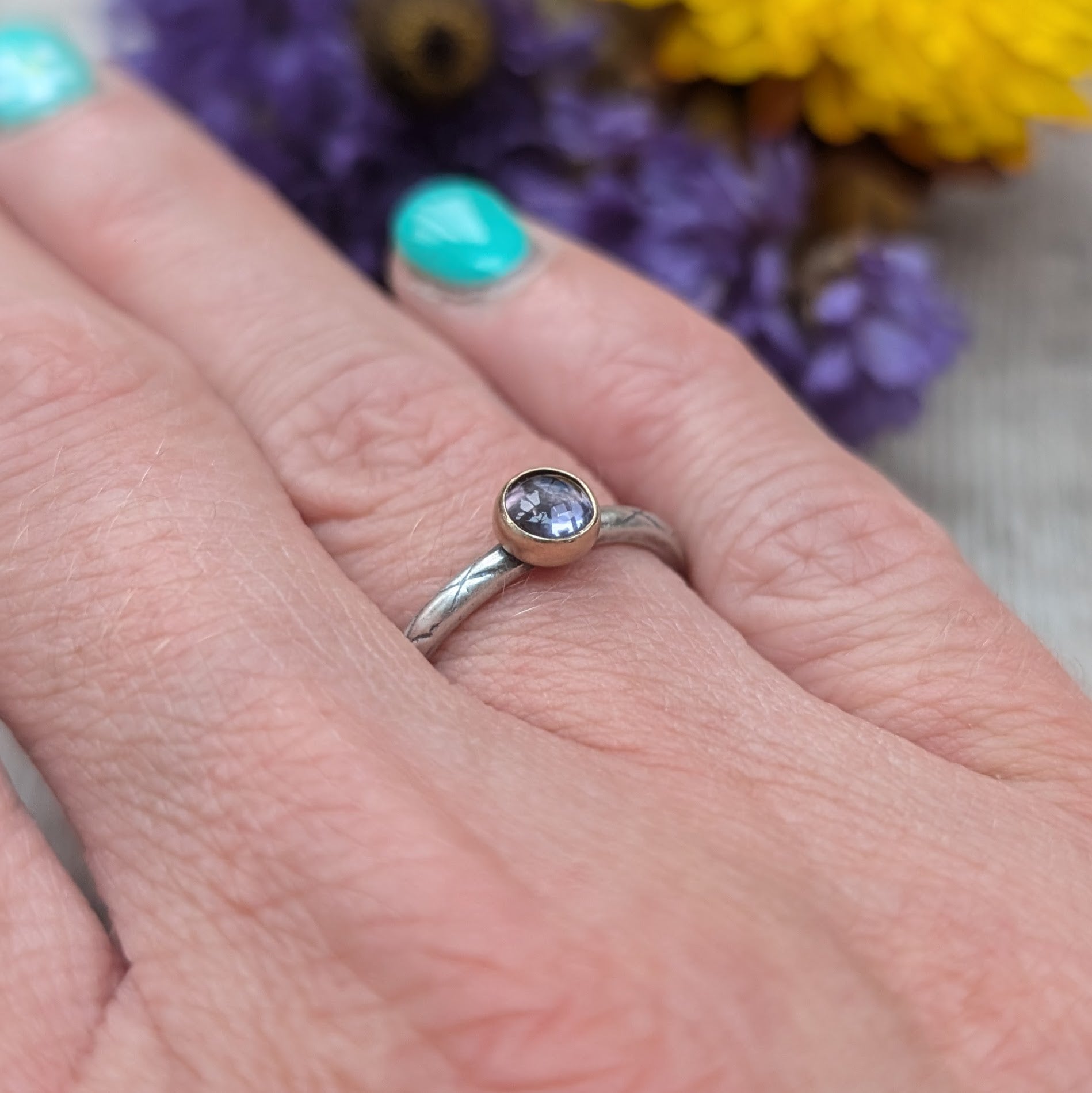 Silver ring with an iolite gemstone set in a gold bezel, worn on a finger. The etched pattern on the band and the deep purple hue of the stone create an elegant and unique look. Background shows blurred flowers in purple and yellow.