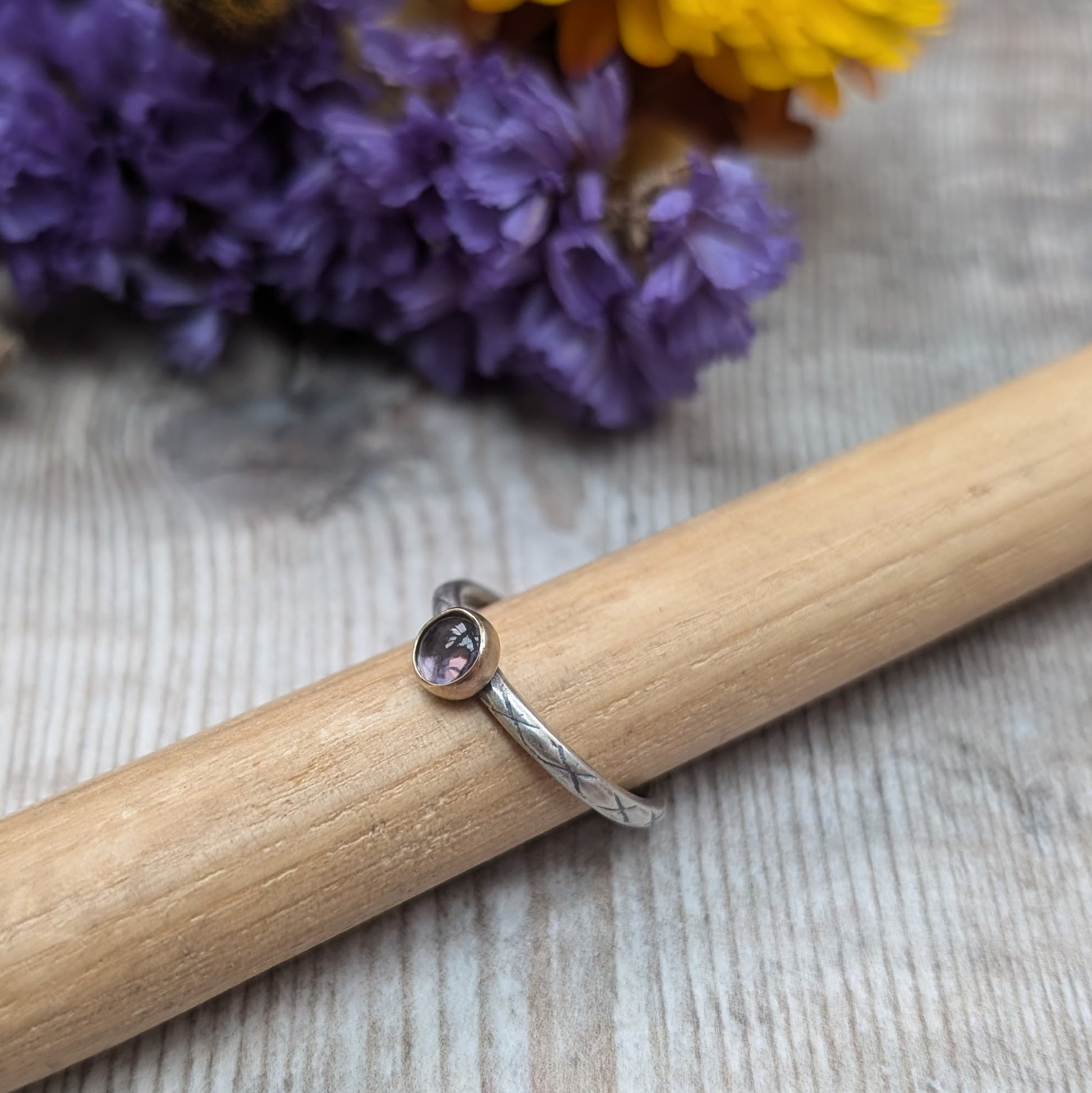 Silver ring with an iolite stone in a gold bezel, displayed on a wooden dowel. The band features an etched pattern, and the background includes purple flowers, adding a natural touch to the setting.