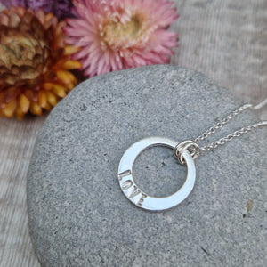 Sterling Silver ‘LOVE’ necklace. Silver disc with offset hole in centre, looks like a thick circle. Attached via two small silver hoops to silver chain. On thicker part of disc, inscription wording hammered into the silver reads LOVE in capitals. Disc measures approximately 20 mm in diameter. Chain length 18 inches.