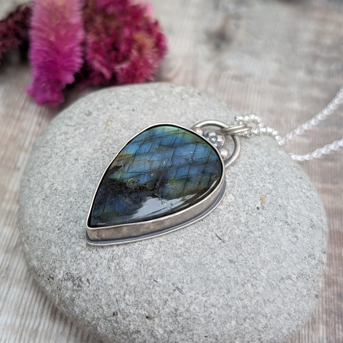 The labradorite pendant displayed on a smooth grey stone, highlighting its shimmering colours. The sterling silver setting features a unique, handcrafted design.
