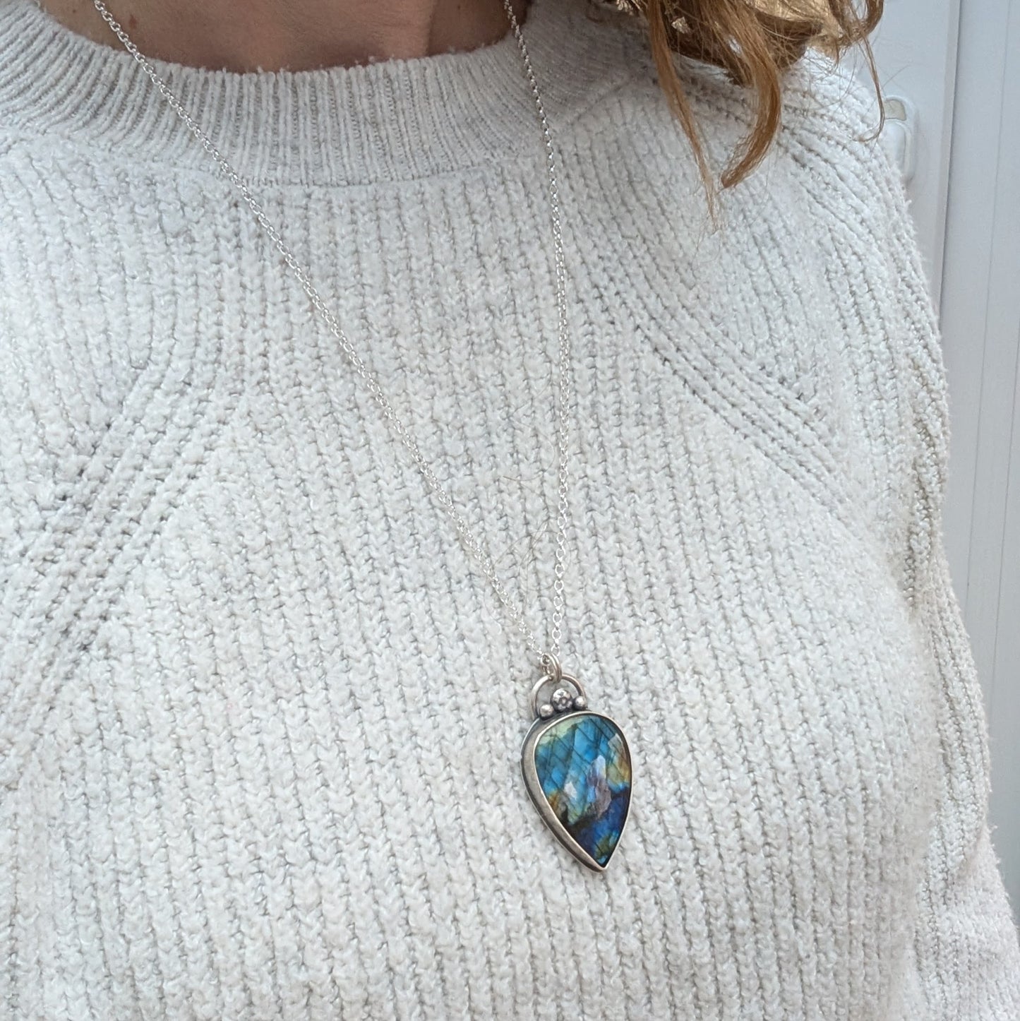 A close-up of a person wearing a long silver necklace with a teardrop-shaped labradorite pendant. The gemstone flashes blue, green, and gold tones, set in recycled sterling silver with decorative silver details.
