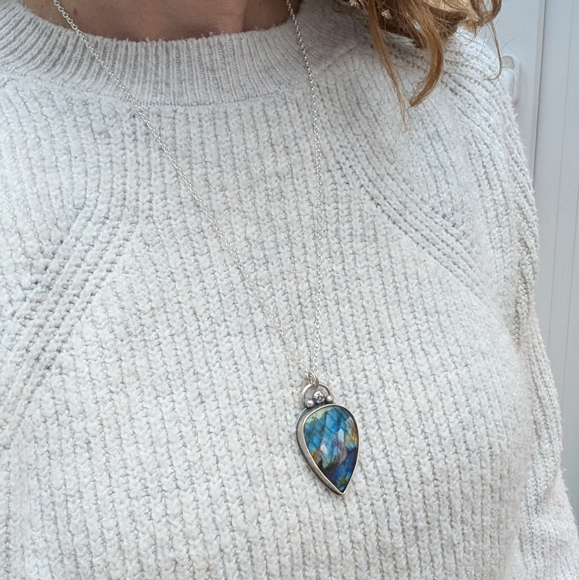 A close-up of a person wearing a long silver necklace with a teardrop-shaped labradorite pendant. The gemstone flashes blue, green, and gold tones, set in recycled sterling silver with decorative silver details.
