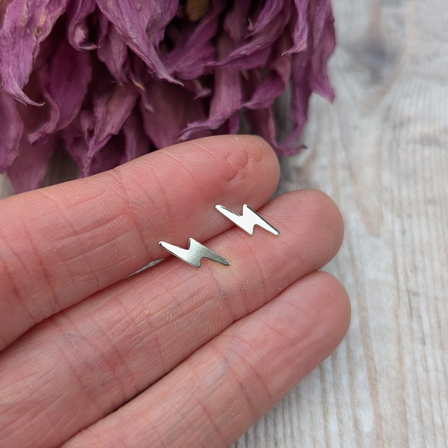 A pair of small silver lightning bolt stud earrings held between fingers, with a dried flower background, highlighting the earrings' fine details.