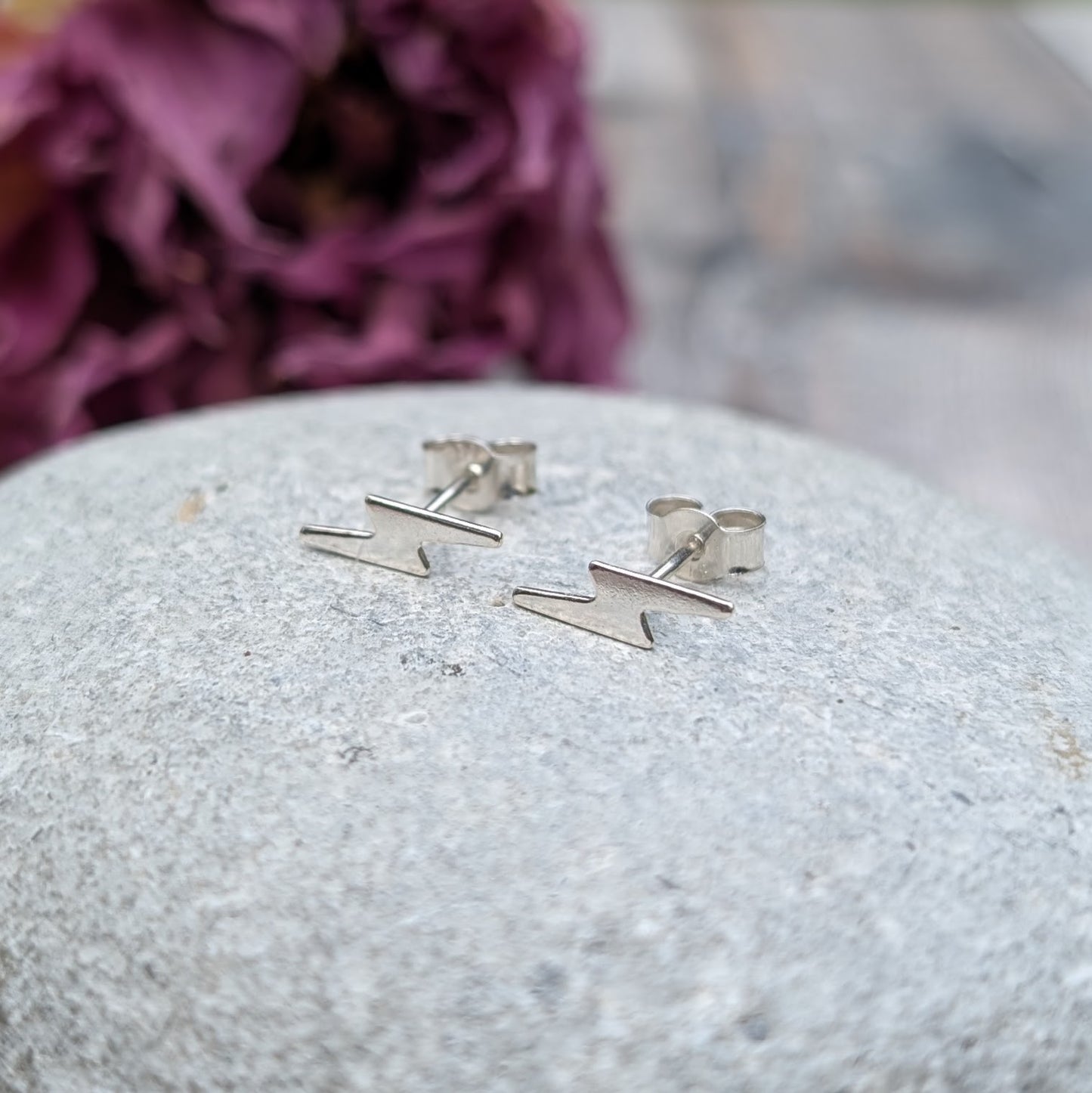 A pair of small silver lightning bolt stud earrings displayed on a smooth stone surface, with a soft purple floral background.