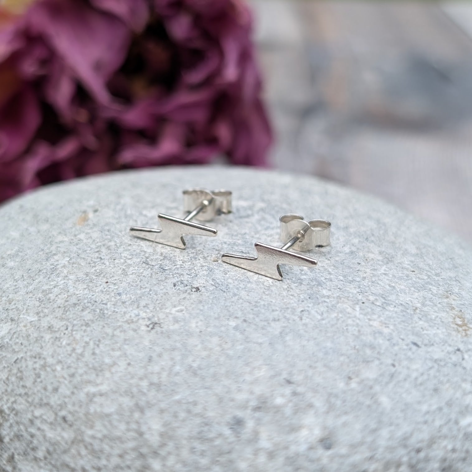 A pair of small silver lightning bolt stud earrings displayed on a smooth stone surface, with a soft purple floral background.