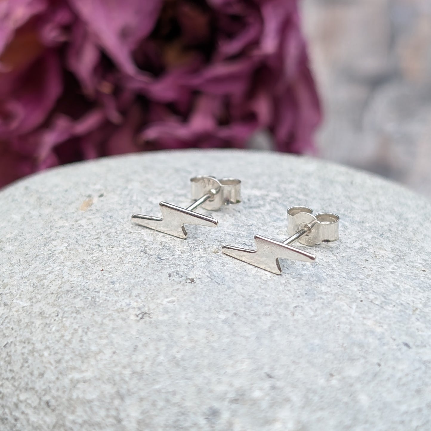 A pair of small silver lightning bolt stud earrings displayed on a smooth stone surface, with a soft purple floral background.