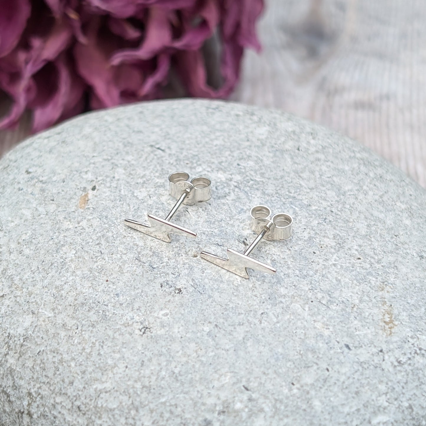 A pair of small silver lightning bolt stud earrings displayed on a smooth stone surface, with a soft purple floral background.