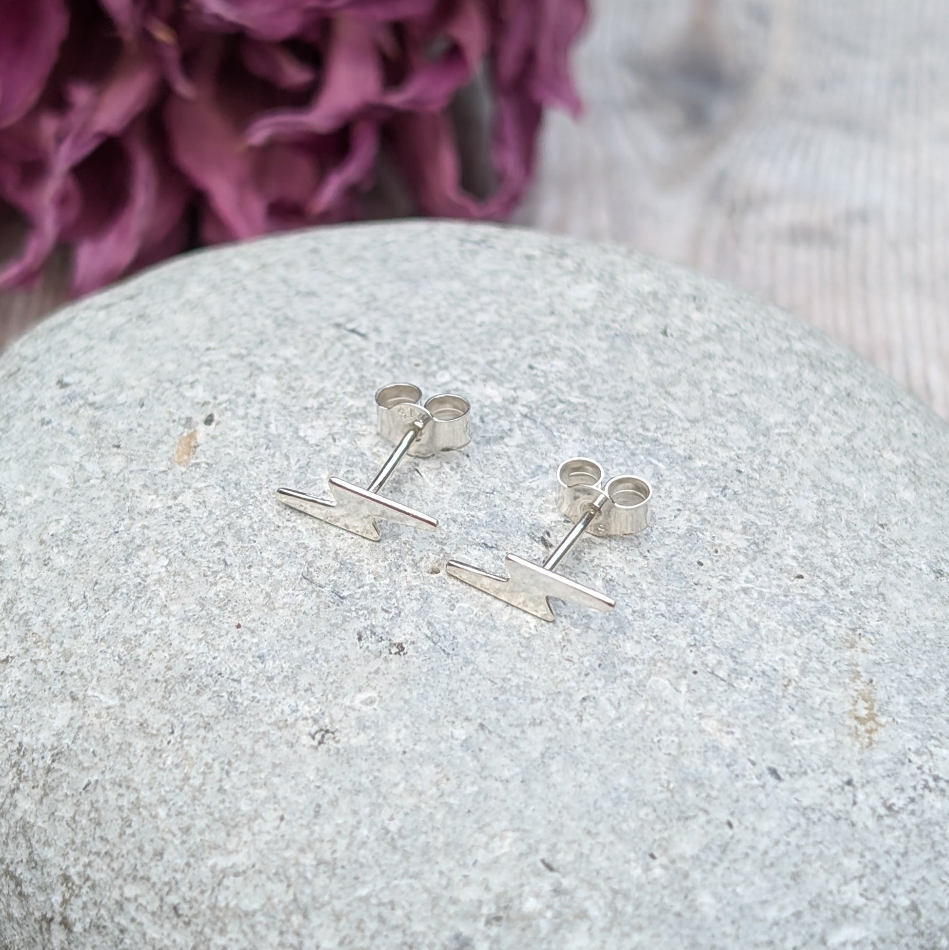 A pair of small silver lightning bolt stud earrings displayed on a smooth stone surface, with a soft purple floral background.
